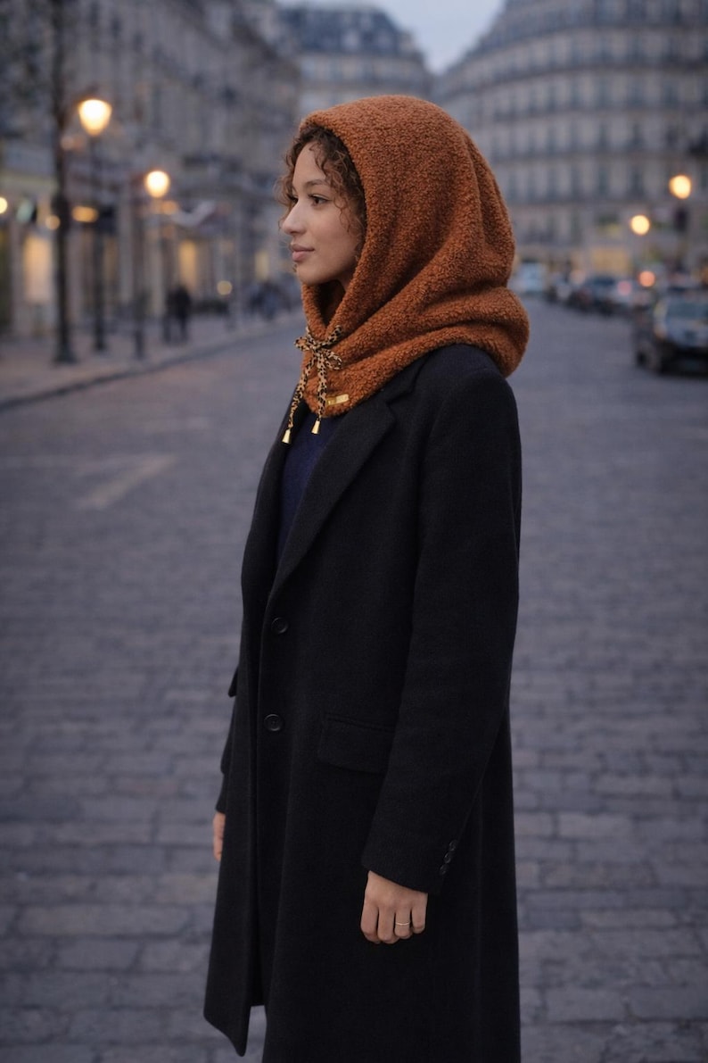 May include: A woman wearing a brown hooded scarf with a decorative tie, paired with a black coat. The scarf is made of a textured material. The background shows a city street with streetlights.