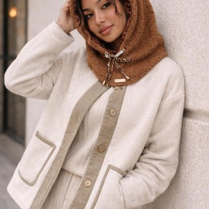 May include: A woman wearing a brown hooded scarf with a cream-colored jacket and matching pants. The jacket has tan trim and pockets. The scarf has a leopard print bow and a small gold tag.