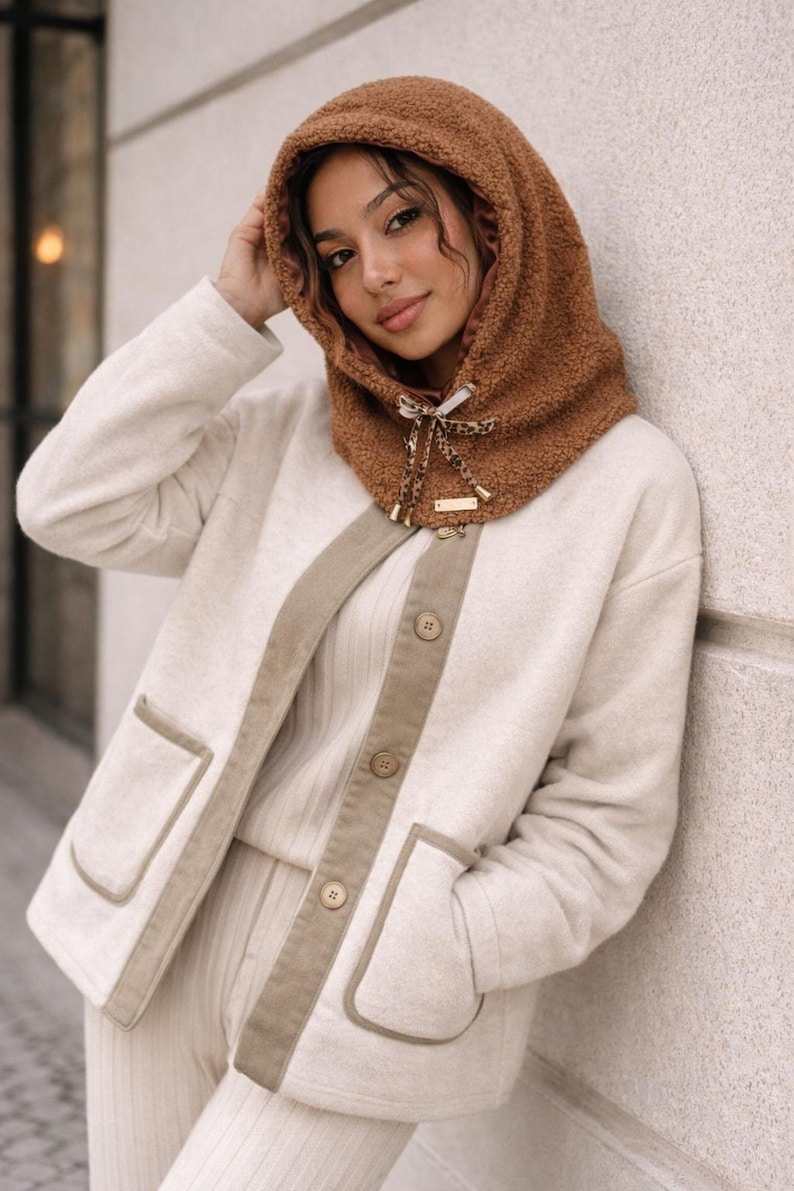 May include: A woman wearing a brown hooded scarf with a cream-colored jacket and matching pants. The jacket has tan trim and pockets. The scarf has a leopard print bow and a small gold tag.