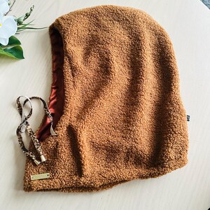 May include: A brown, textured balaclava with a satin lining. The hood features a drawstring with a leopard print pattern and a small gold-colored tag. The balaclava is designed to cover the head and neck.