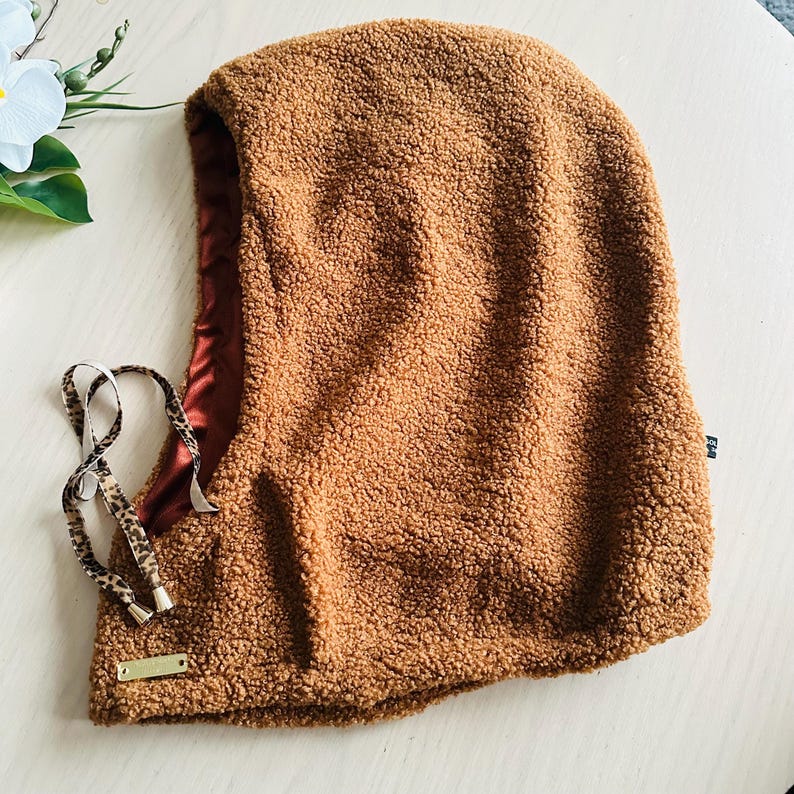 May include: A brown, textured balaclava with a satin lining. The hood features a drawstring with a leopard print pattern and a small gold-colored tag. The balaclava is designed to cover the head and neck.