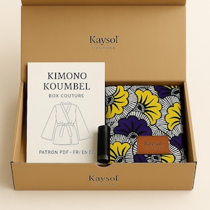 Op de afbeelding: Een bruine kartonnen doos met een naaiset. Binnenin een patroonboek met de tekst "KIMONO KOUMBEL BOX COUTURE" en een stof met gele en paarse bloemmotieven. Een zwart cilindrisch object en een leren label zijn ook zichtbaar.