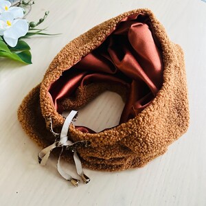 May include: A brown, fluffy neck warmer with a satin lining. The neck warmer has a drawstring closure with metallic accents. The interior is a deep reddish-brown color. The outer material has a textured, plush appearance.