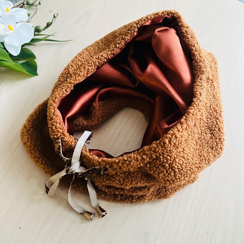 May include: A brown, fluffy neck warmer with a satin lining. The neck warmer has a drawstring closure with metallic accents. The interior is a deep reddish-brown color. The outer material has a textured, plush appearance.