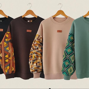 Könnte beinhalten: Vier Sweatshirts mit Rundhalsausschnitt in Schwarz, Braun, Beige und Türkis. Jedes hat einen einfarbigen Körper und gemusterte Ärmel mit geometrischen Designs. Auf der Brust befindet sich ein kleines rechteckiges Patch. Sie hängen an Holzbügeln.