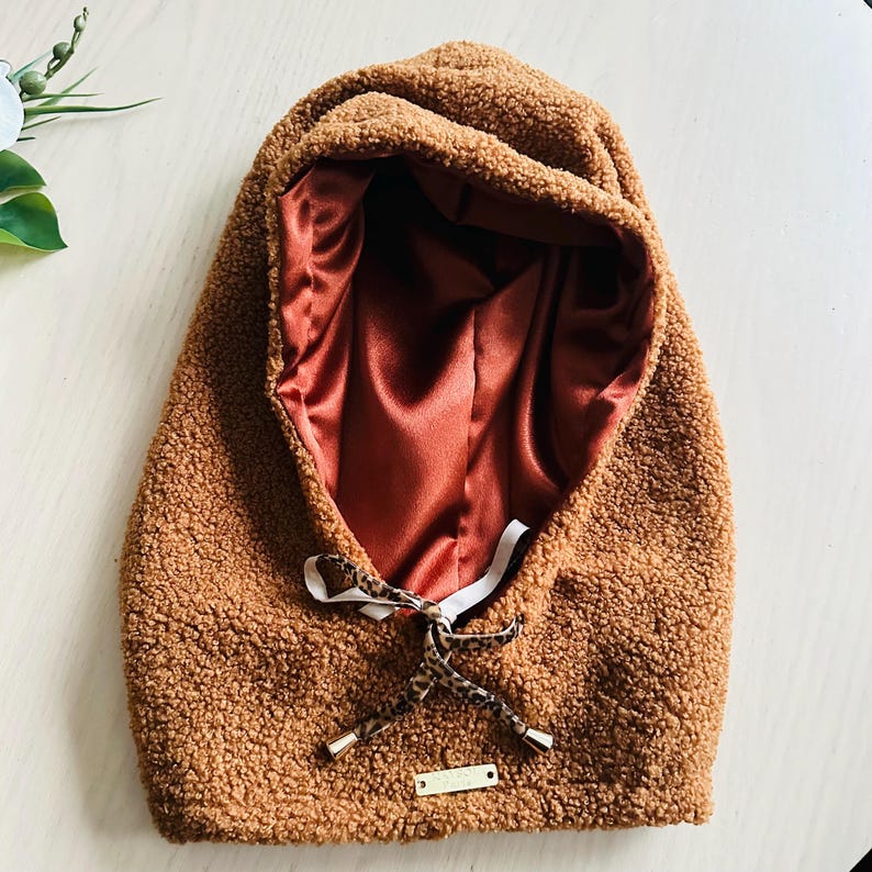 May include: A brown, hooded winter hat with a soft, textured exterior. The interior is lined with a rust-colored satin fabric. A drawstring with a leopard print pattern and gold-colored aglets is tied at the front. A small, gold-colored tag is attached.