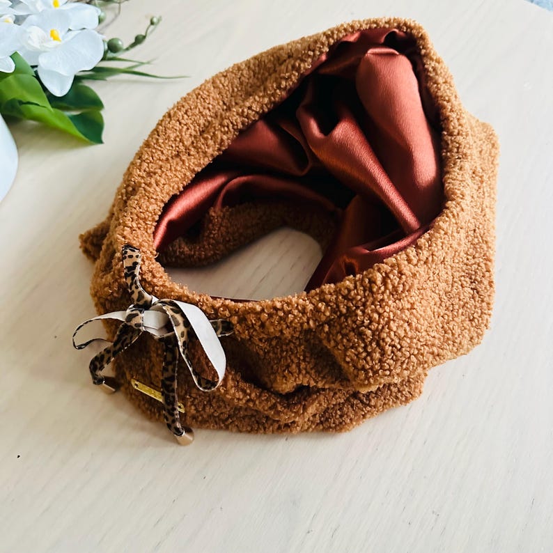 May include: A brown, textured neck warmer with a satin lining. The neck warmer has a leopard print bow and a white ribbon. The interior is a reddish-brown color. The neck warmer is on a light-colored surface.