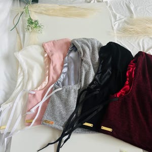 Puede incluir: Una colección de gorros en varios colores, incluyendo blanco, rosa, gris, negro y burdeos. Cada gorro presenta un forro de satén y un cierre de cordón. Pequeñas etiquetas rectangulares doradas están adjuntas.