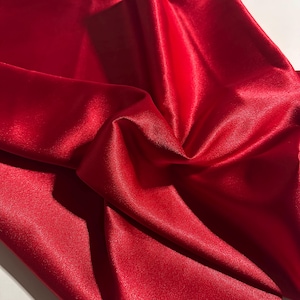 Op de afbeelding: Close-up van een levendige rode satijnen stof. De stof is gedrapeerd en gevouwen, waardoor schaduwen en highlights ontstaan die de gladde textuur en glans benadrukken. Het materiaal ziet er luxueus uit.