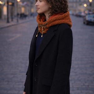 May include: A person wearing a black coat and a rust-colored hooded scarf with a decorative tie. The scarf has a soft, textured appearance. The background shows a city street with buildings and streetlights.