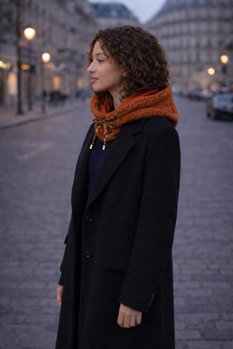 May include: A person wearing a black coat and a rust-colored hooded scarf with a decorative tie. The scarf has a soft, textured appearance. The background shows a city street with buildings and streetlights.