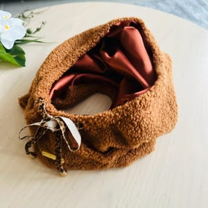 May include: A brown, fluffy, circular scarf with a satin interior. The scarf has a leopard print bow with a gold-colored tag. The interior is a reddish-brown color. The scarf is on a white surface.