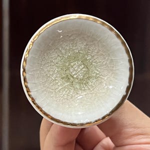 Puede incluir: Un pequeño cuenco redondo de cerámica blanca con borde dorado. El cuenco tiene un patrón de esmalte craquelado y un centro ligeramente elevado. El cuenco está sostenido en la mano de una persona.