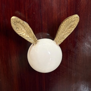 May include: A white ceramic rabbit head with gold leaf ears. The rabbit head is a decorative piece and is mounted on a dark wood surface.