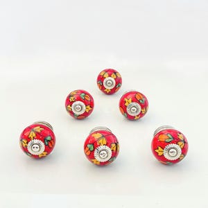 May include: Six red ceramic cabinet knobs with a floral design in yellow, green, and black. The knobs have a silver metal base.