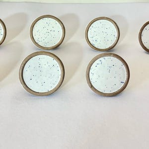 May include: Eight ceramic cabinet knobs. Each knob has a white speckled face with blue dots, a brown rim, and a brown base. The knobs are arranged in two rows of four on a light background.