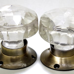 Può includere: Due pomelli per porte in vetro trasparente con bordi sfaccettati e basi in ottone. I pomelli sono rotondi e hanno una finitura lucida.