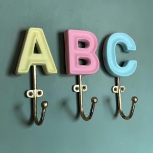 May include: Three wall-mounted hooks featuring the letters A, B, and C. The letter A is pale yellow, B is light pink, and C is light blue. Each letter is attached to a metallic gold-toned hook with two screw holes for mounting to a flat surface.