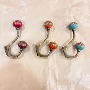 Peut inclure: Trois patères murales décoratives avec des bras en métal couleur laiton et des boutons en céramique colorés. Les boutons sont rouges, oranges et turquoise, avec des motifs complexes. Les crochets sont conçus pour suspendre des objets.