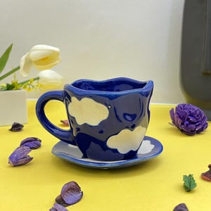 May include: A blue ceramic teacup with a white cloud design. The teacup has a wavy rim and a matching saucer.