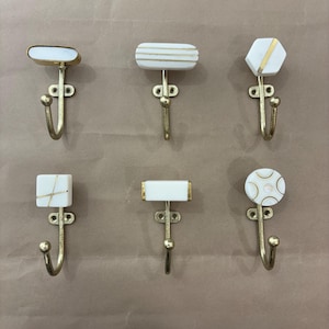 May include: Six decorative wall hooks with white and gold detailing. Each hook features a unique design on the white top, such as stripes, geometric shapes, and a circular pattern. The hooks are made of gold-coloured metal.