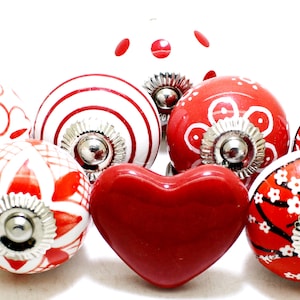 Peut inclure: Un ensemble de huit boutons de tiroir en céramique rouge et blanche avec un bouton en forme de cœur au centre. Les boutons présentent différents motifs, notamment des pois, des rayures et des motifs floraux.