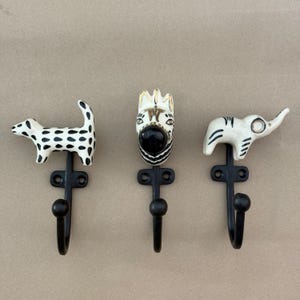 Könnte beinhalten: Drei dekorative Wandhaken mit Tiermotiven: ein Dalmatiner, ein Zebra und ein Elefant. Jeder Haken hat eine schwarze Metallbasis und einen gebogenen Haken. Die Tierfiguren sind weiß mit schwarzen Markierungen.