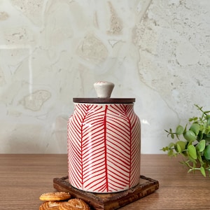 Puede incluir: Tarro de cerámica blanco con un patrón de chevron rojo y blanco y una tapa de madera. El tarro está sentado en una posavasos de madera con algunas galletas en forma de corazón al lado.