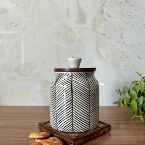 Op de afbeelding: Witte keramische pot met een zwart-wit chevronpatroon. De pot heeft een houten deksel met een knop. De pot staat op een houten onderzetter met koekjes ernaast.