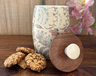 May include: A ceramic cookie jar with a floral pattern in shades of blue, pink, and green. The jar has a wooden lid with a white knob.