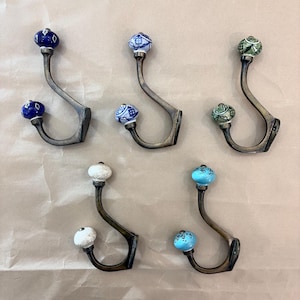 May include: Set of five decorative metal wall hooks with ceramic knobs. The hooks have a curved design and are in a bronze finish. The ceramic knobs are in various colors, including blue, white, and green, with patterned designs.