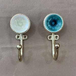 May include: Two decorative wall hooks. One hook has a white and gold design, the other has a teal and gold design. Both hooks have a gold-coloured metal base with a curved hook and a decorative top.