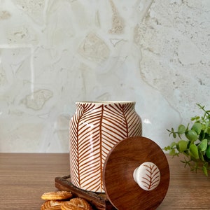 Puede incluir: Un tarro de cerámica blanco con un patrón de hojas marrones y una tapa de madera. La tapa tiene un pomo blanco con un diseño de hoja. Hay galletas en una bandeja de madera junto al tarro.