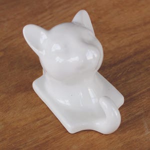 May include: White ceramic cat head wall hook. The cat's head is facing to the right and the tail is curled into a hook.