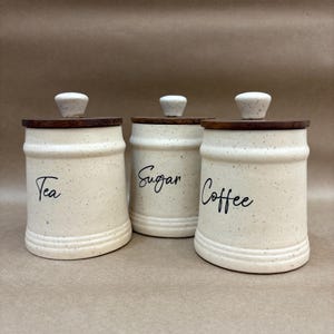 May include: Three ceramic storage jars with wooden lids. The jars are off-white with black speckles and labelled "Tea", "Sugar", and "Coffee" in a handwritten font. Each jar has a small, off-white knob on top.