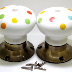 Könnte beinhalten: Zwei weiße Keramik-Türknöpfe mit Messingsockel. Die Knöpfe sind mit bunten Punkten verziert.