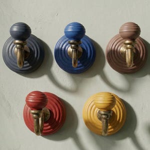 May include: A set of five wall hooks featuring circular, ribbed backplates in dark grey, blue, brown, red, and yellow. Each hook includes a matching spherical knob and a curved, antiqued brass-toned metal arm extending from the centre.