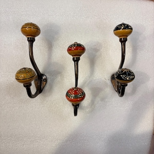 Puede incluir: Tres percheros de pared decorativos con pomos de cerámica ornamentados en tonos amarillos, rojos y negros. Los ganchos tienen un acabado bronce y están montados en una pared blanca. Los pomos presentan diseños florales.