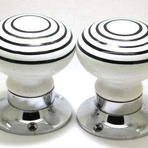 May include: Pair of white ceramic door knobs with black concentric circle design. The knobs have a rounded shape and are mounted on a chrome-plated base. Decorative hardware for doors.