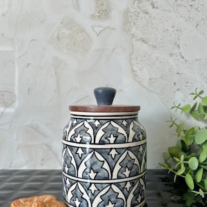 Op de afbeelding: Een keramische pot met een houten deksel en een donkergrijze knop. De pot is versierd met een herhalend patroon van witte en grijze vormen. De pot staat op een zwart oppervlak naast hartvormige koekjes.