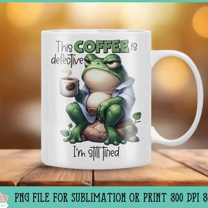 May include: White ceramic coffee mug featuring a green frog illustration holding a cup of coffee. The mug has the text "This coffee is defective I'm still tired" printed on it.