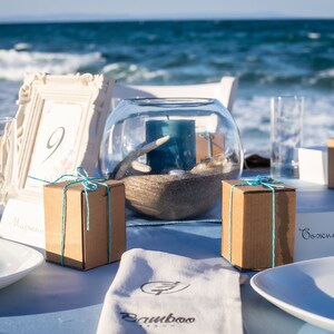 May include: A table setting with a white tablecloth, two white plates, two glasses, two small brown boxes tied with blue ribbon, a white picture frame with the number 9, a glass bowl with sand, a blue candle, and a white napkin with the text "Bamboo" printed on it.