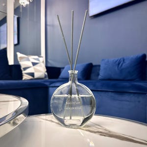 Op de afbeelding: Een glazen diffuserfles met rietjes in een transparante kleur met grijze rietjes. De fles heeft de tekst "TRÅKE Reed Diffuser | PREMIUM BLEND" aan de voorkant.