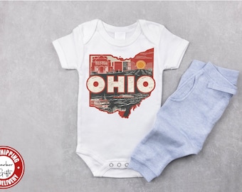 May include: White baby onesie with a vintage-style illustration of the state of Ohio, featuring landmarks and the word "OHIO" in red letters. The onesie is paired with light blue baby pants.