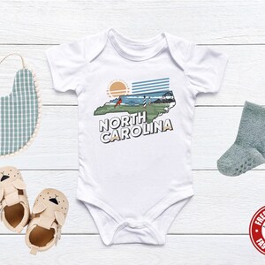 May include: White baby bodysuit with a graphic of the state of North Carolina, a lighthouse, and the text "North Carolina".