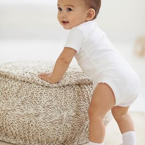 May include: A baby in a white onesie and white socks is standing next to a beige knitted ottoman.