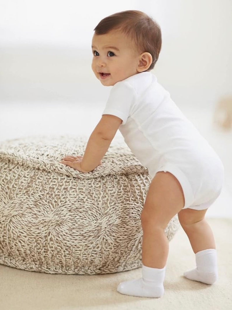 May include: A baby in a white onesie and white socks is standing next to a beige knitted ottoman.