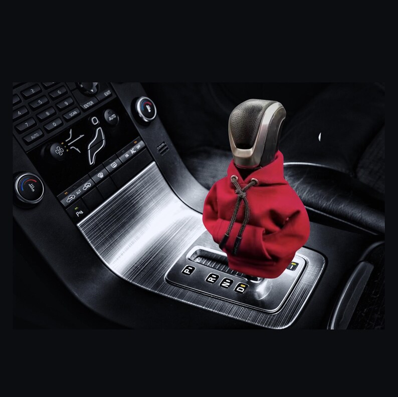 May include: A red hooded sweatshirt with a silver head and face covering is attached to a car's gear shift lever. The gear shift lever has the letters P, R, N, D, and a number 1.