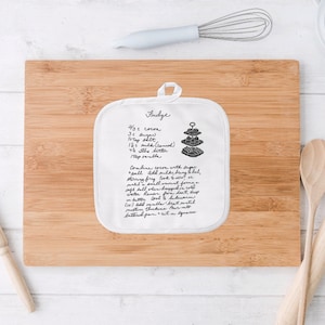 May include: White cotton pot holder with a handwritten fudge recipe. The recipe includes ingredients like cocoa, sugar, milk, and butter. The pot holder is on a wooden cutting board.