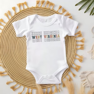 May include: White baby onesie with the text "WEST VIRGINIA" repeated three times in a stacked, retro-style font with colorful outlines. The onesie is on a woven straw mat with fringe.